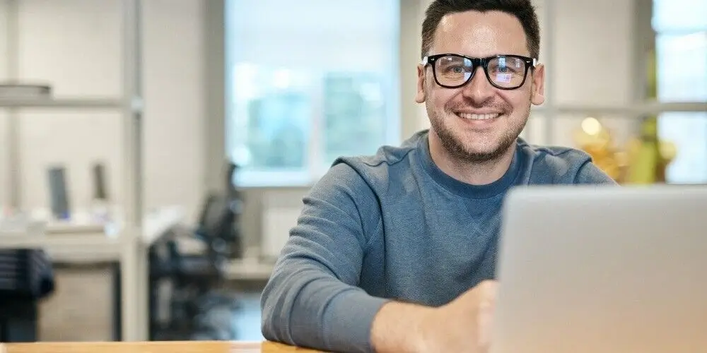 A smiling man wearing glasses and a blue sweater working on a laptop in a bright office setting.