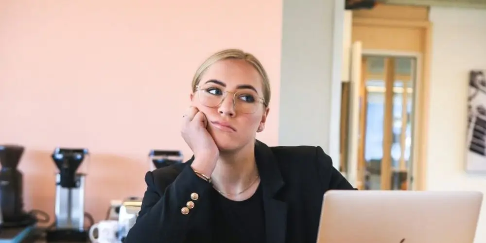 A woman in a black blazer and glasses sits at a desk, resting her head on her hand, looking thoughtful or bored, with a laptop in front of her. The office setting includes coffee machines and modern decor.