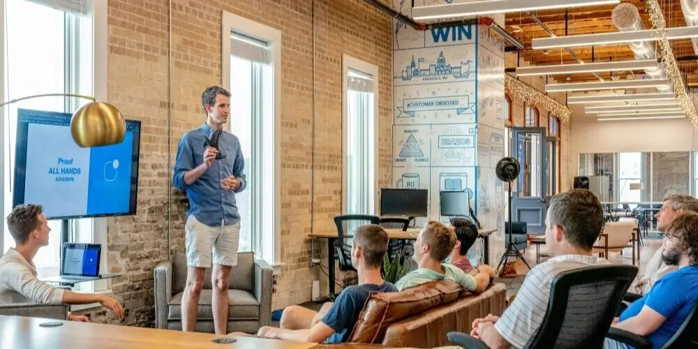 Presenter giving a talk to a group of people seated in a modern office environment, with a large screen displaying a presentation.