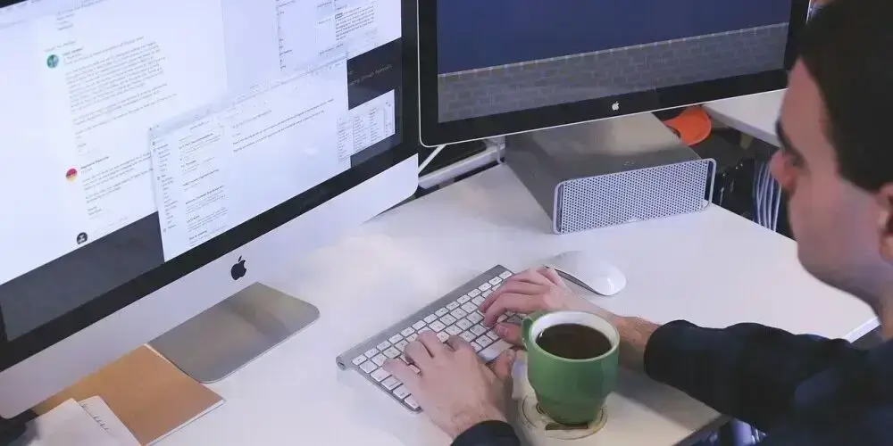 A person working on a dual-monitor setup while holding a cup of coffee, representing long hours of work at a desk in a modern office environment.