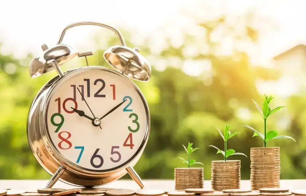 An alarm clock next to three stacks of coins with small plants growing on top, symbolizing the concept of time and financial growth.