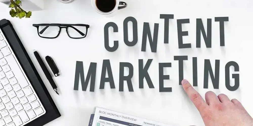 A workspace featuring the phrase 'Content Marketing' with a keyboard, glasses, pen, coffee, and a hand pointing to the word 'Marketing.