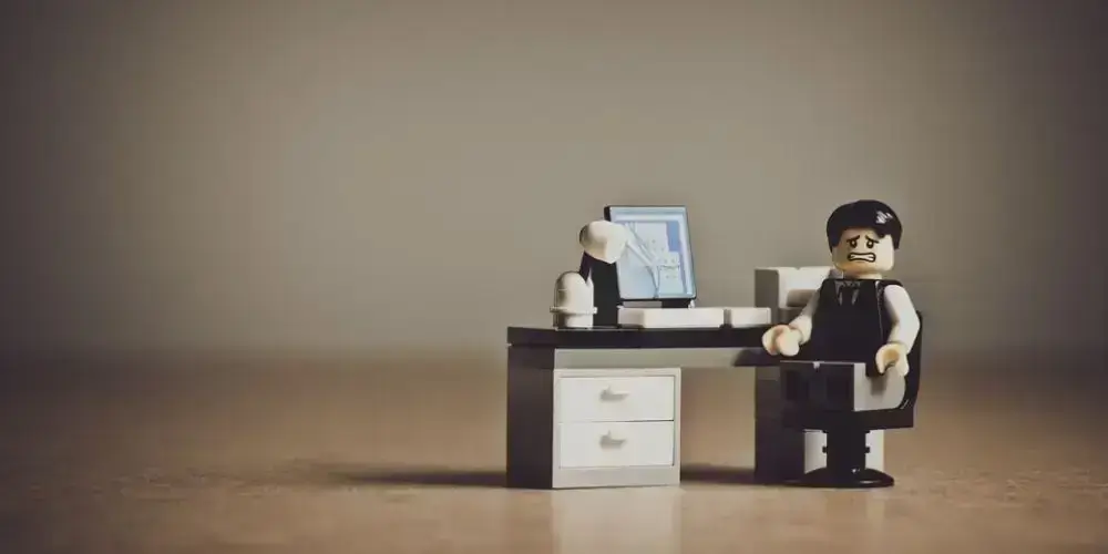 A LEGO figurine looking stressed while sitting at a desk with a computer, depicting the stress of sedentary work.