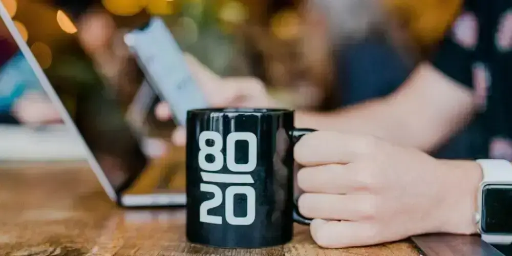 A person holding a coffee mug labeled "80/20" while working on a laptop, symbolizing the application of the 80/20 rule in productivity and affiliate marketing.