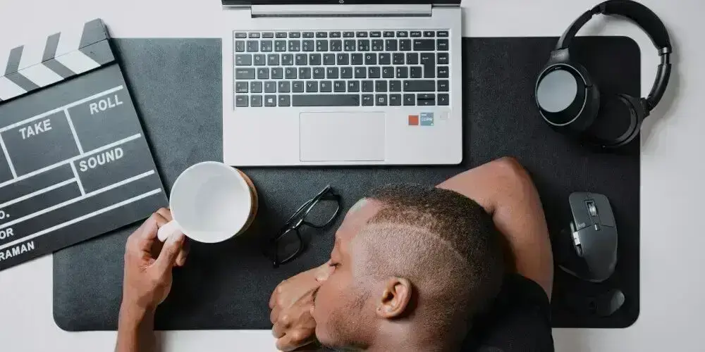 A person resting their head on a desk beside a laptop and coffee, symbolizing the effects of procrastination and lack of motivation in affiliate marketing.